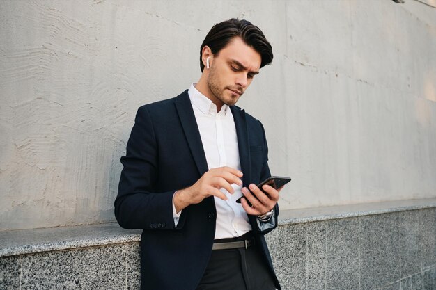 Jeune homme brune barbu sérieux séduisant en chemise blanche et costume classique avec des écouteurs sans fil utilisant judicieusement un téléphone portable debout près du mur dans la rue seule