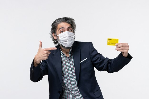 Jeune homme de bureau en costume portant un masque et montrant sa carte bancaire pointant quelque chose sur le côté gauche sur fond blanc