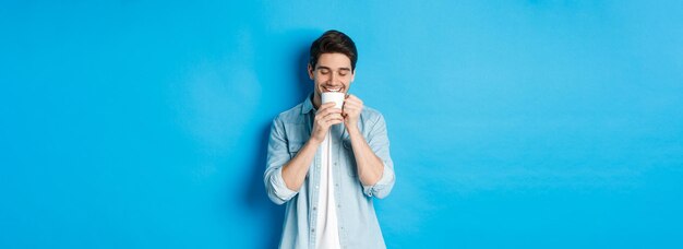 Jeune homme buvant du café avec un visage heureux debout sur fond bleu