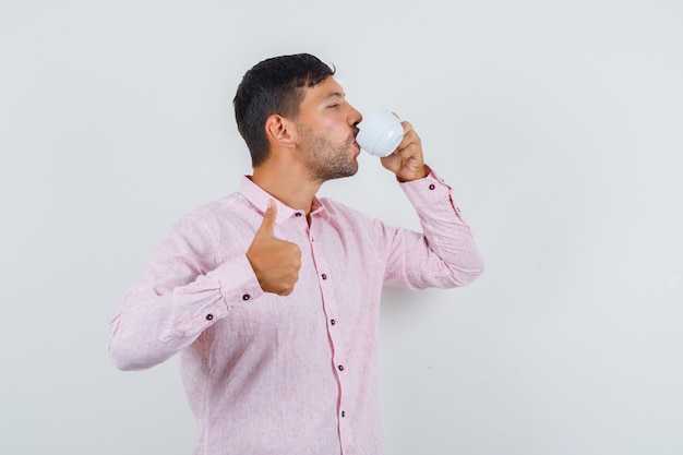 Jeune homme buvant du thé avec le pouce vers le haut en vue de face de chemise rose.