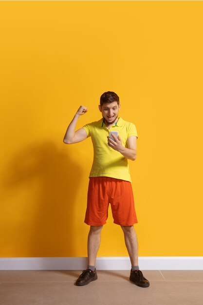 Jeune homme caucasien à l'aide de smartphone. Portrait de toute la longueur du corps isolé sur mur jaune.