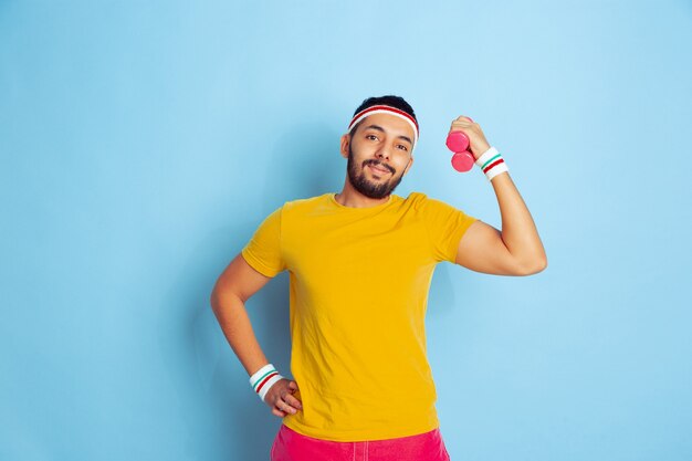 Jeune homme caucasien en formation de vêtements lumineux sur fond bleu Concept de sport, émotions humaines, expression faciale, mode de vie sain, jeunesse, ventes. Entraînement avec les poids colorés. Copyspace.