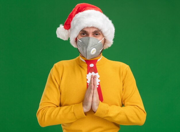 Jeune homme caucasien portant chapeau de Noël et cravate avec masque de protection regardant la caméra en gardant les mains ensemble isolé sur fond vert