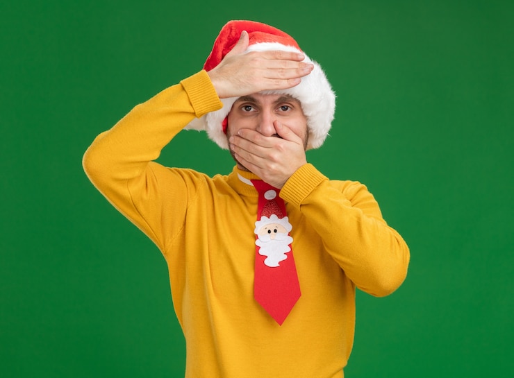 Photo gratuit jeune homme caucasien portant chapeau de noël et cravate regardant la caméra en gardant les mains sur la bouche et sur le front isolé sur fond vert