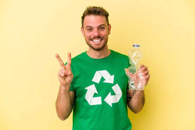 Jeune homme caucasien tenant une bouteille de plastique à recycler isolé sur fond jaune montrant le numéro deux avec les doigts.