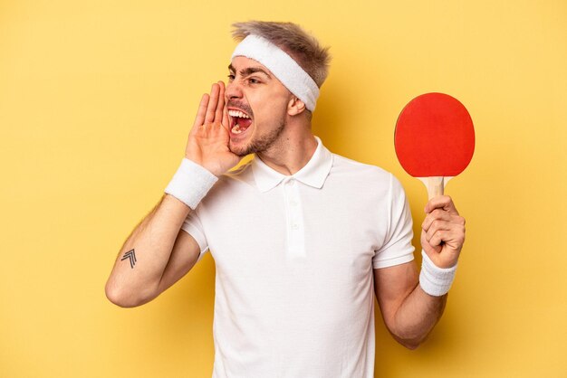 Jeune homme caucasien tenant des raquettes de ping-pong isolées sur fond jaune criant et tenant la paume près de la bouche ouverte.
