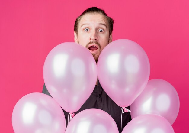 Jeune homme célébrant la fête d'anniversaire furtivement sur les ballons étonné et surpris debout sur le mur rose
