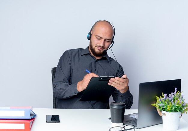 Jeune homme de centre d'appels chauve concentré portant un casque assis au bureau avec des outils de travail écrit avec un stylo sur le presse-papiers isolé sur blanc