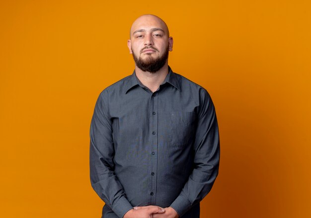 Jeune homme de centre d'appels chauve debout et regardant la caméra et gardant les mains ensemble isolé sur fond orange avec espace de copie