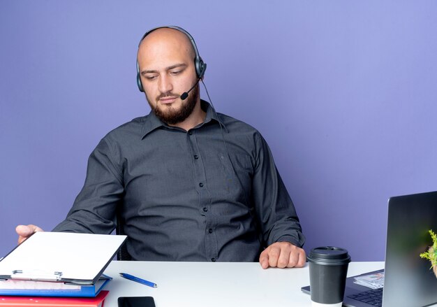 Jeune homme de centre d'appels chauve portant un casque assis au bureau avec des outils de travail touchant et regardant le presse-papiers isolé sur fond violet