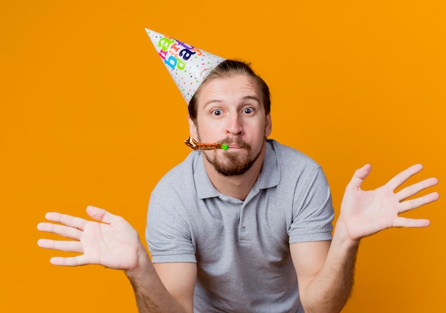 Jeune homme en chapeau de fête soufflant un sifflet heureux et surpris de répandre les bras sur les côtés concept de fête d'anniversaire debout sur un mur orange