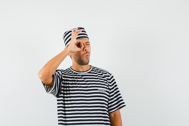 Jeune homme en chapeau t-shirt rayé montrant le geste ok sur les yeux et à la curiosité