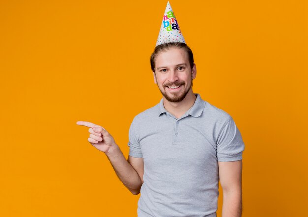 Jeune homme avec chapeau de vacances célébrant la fête d'anniversaire heureux et gai souriant pointant avec le doigt sur le côté debout sur le mur orange