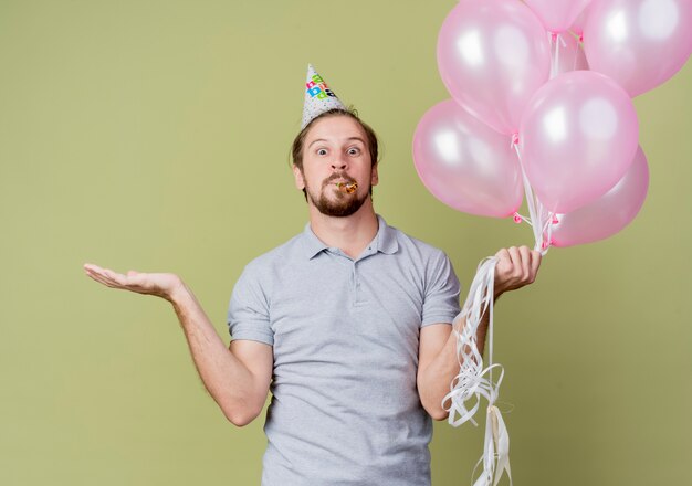 Jeune homme avec chapeau de vacances tenant des ballons à la fête d'anniversaire de célébration confus debout sur un mur léger