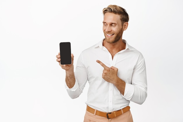 Jeune homme charismatique pointant vers un smartphone et souriant démontrant une publicité sur fond blanc de téléphone portable