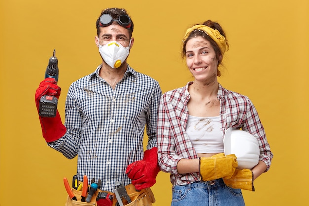 Jeune homme charpentier portant des lunettes de protection et un masque tenant la machine de forage étant équipé de différents outils pour la construction debout près de sa femme ayant une expression heureuse tout en travaillant