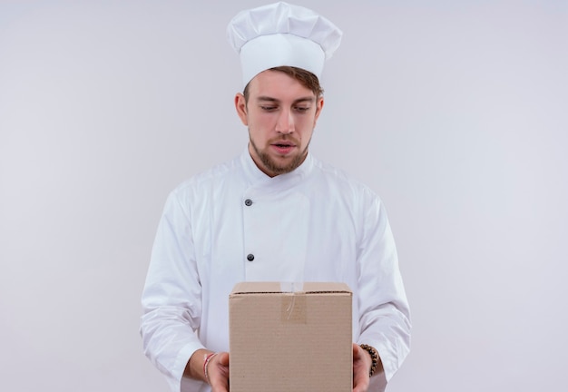 Un jeune homme chef barbu surpris en uniforme blanc tenant la boîte de livraison et en le regardant sur un mur blanc