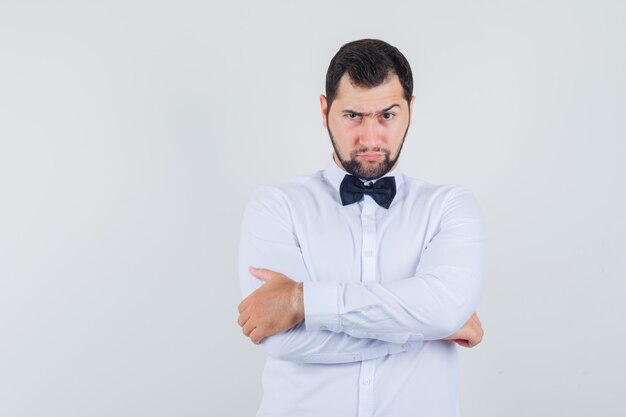 Jeune homme en chemise blanche debout avec les bras croisés et à la grave, vue de face.