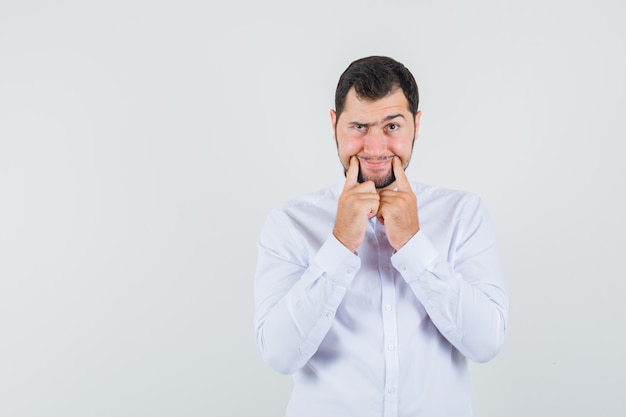 Jeune homme en chemise blanche forçant un sourire sur le visage et regardant sombre, vue de face.