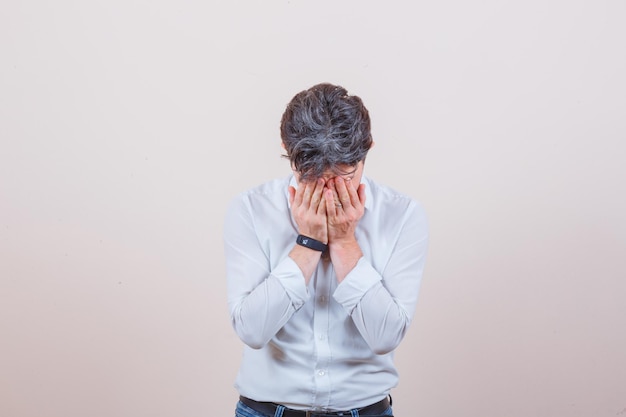 Jeune homme en chemise blanche, jeans couvrant le visage avec les mains et à la colère