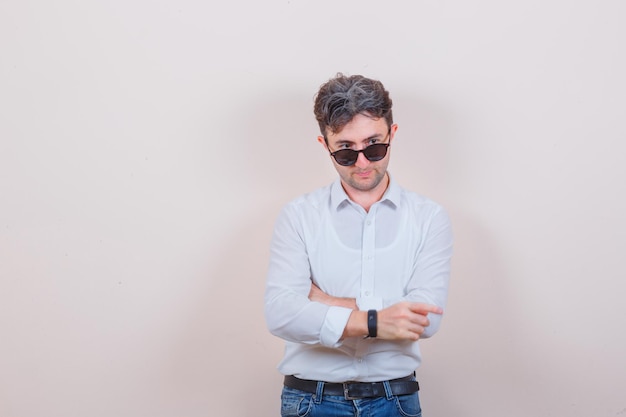 Jeune homme en chemise blanche, jeans posant tout en regardant par-dessus des lunettes et l'air pensif