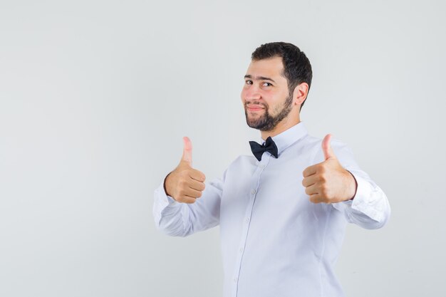 Jeune homme en chemise blanche montrant deux pouces vers le haut et à la joyeuse vue de face.