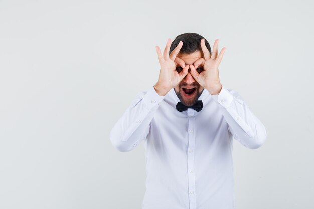 Jeune homme en chemise blanche montrant le geste de lunettes et regardant étonné, vue de face.