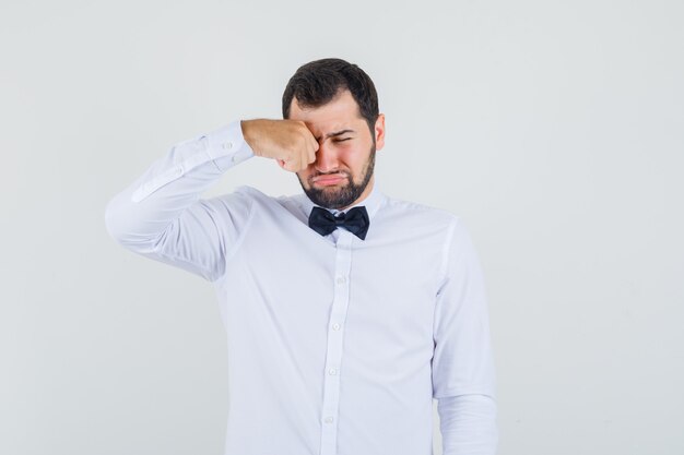 Jeune homme en chemise blanche se frottant les yeux en pleurant, vue de face.