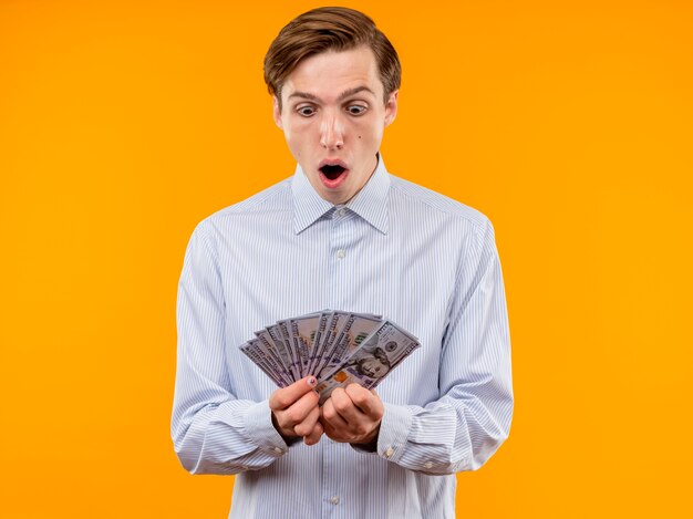 Jeune homme en chemise blanche tenant de l'argent à la surprise et surpris debout sur un mur orange