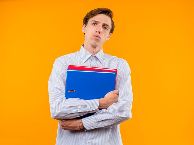 Jeune homme en chemise blanche tenant des dossiers à la recherche avec l'expression confiante serioua debout sur un mur orange