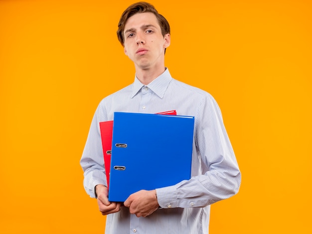 Jeune homme en chemise blanche tenant des dossiers avec un visage sérieux debout sur un mur orange