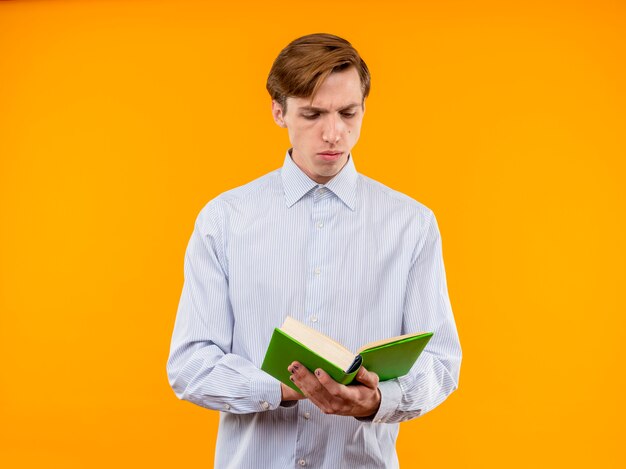 Jeune homme en chemise blanche tenant un livre ouvert en le regardant avec un visage sérieux debout sur un mur orange