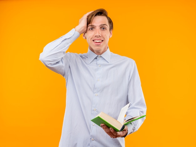 Jeune homme en chemise blanche tenant un livre ouvert à sourire joyeusement surpris debout sur un mur orange