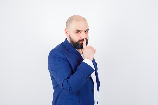 Jeune homme en chemise blanche, veste montrant un geste de silence et l'air confiant, vue de face.