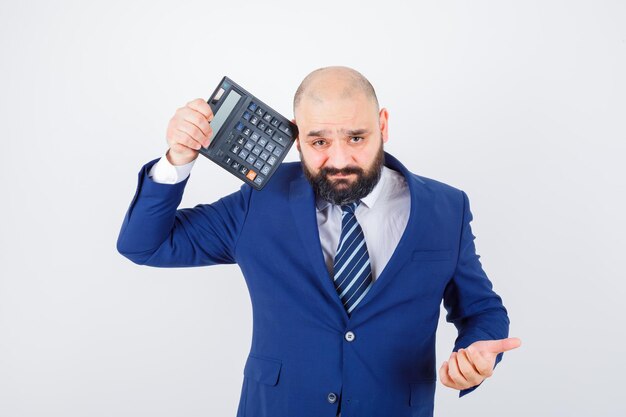 Jeune homme en chemise blanche, veste tenant une calculatrice près de l'œil et l'air confus, vue de face.