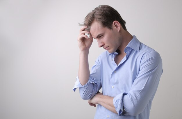 Jeune homme avec une chemise bleue ayant des pensées profondes debout contre un mur blanc