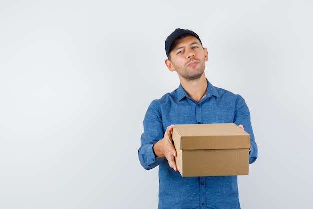 Jeune homme en chemise bleue, casquette présentant une boîte en carton, vue de face.