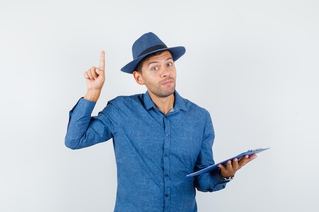 Jeune homme en chemise bleue, chapeau tenant le presse-papiers avec le doigt vers le haut et l'air curieux, vue de face.