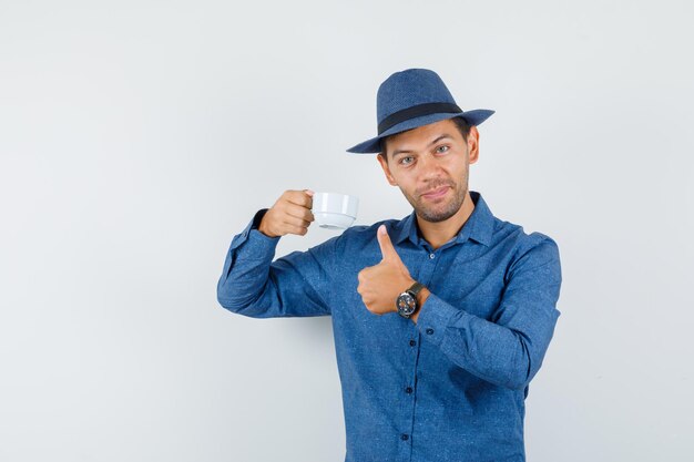 Jeune homme en chemise bleue, chapeau tenant une tasse de boisson avec le pouce vers le haut et l'air joyeux, vue de face.