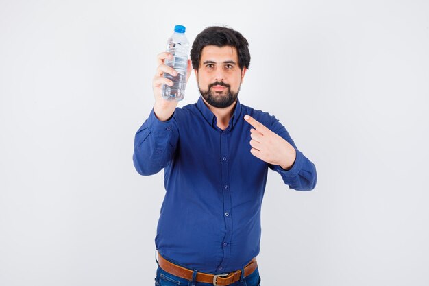 Jeune homme en chemise bleue et jeans tenant une bouteille d'eau et la pointant et l'air optimiste, vue de face.
