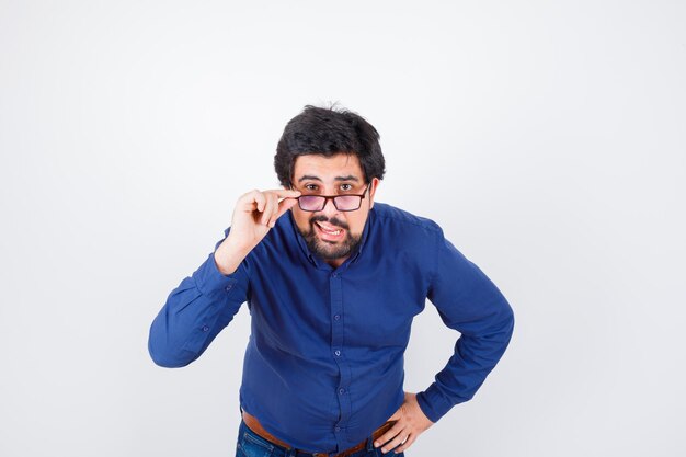 Jeune homme en chemise bleue et jeans tenant la main sur la taille tout en enlevant les lunettes et l'air curieux, vue de face.