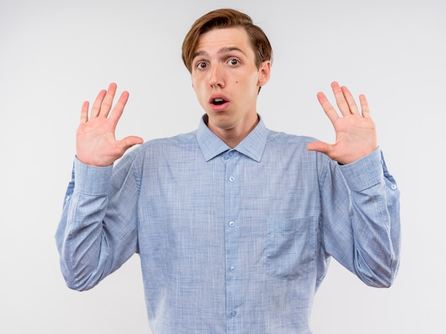 Jeune homme en chemise bleue en levant les mains dans l'abandon d'avoir peur debout sur fond blanc