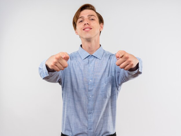 Jeune homme en chemise bleue pointant avec l'index souriant confiant heureux et positif debout sur un mur blanc