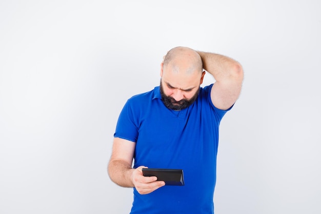 Jeune homme en chemise bleue regardant la calculatrice tout en se grattant la tête et l'air perdu, vue de face.