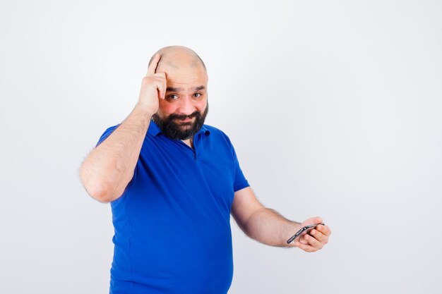 Jeune homme en chemise bleue regardant le téléphone tout en pointant vers sa tête et en ayant l'air plein d'espoir, vue de face.