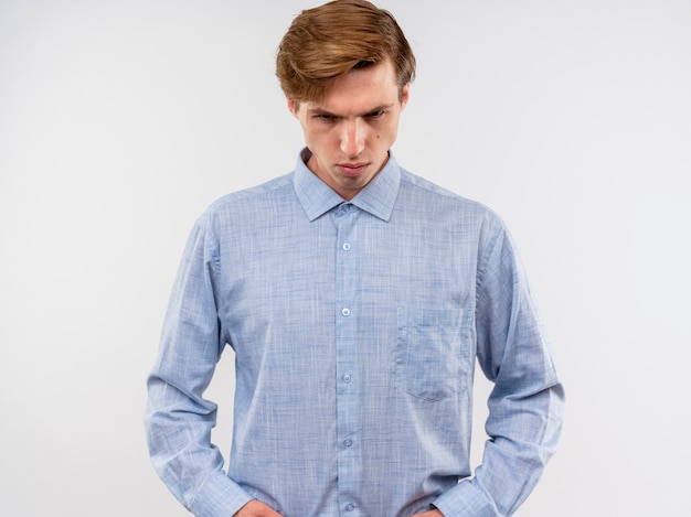 Photo gratuite jeune homme en chemise bleue regardant vers le bas avec une expression sérieuse debout sur un mur blanc