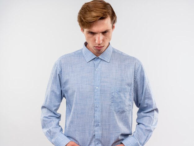 Jeune homme en chemise bleue regardant vers le bas avec une expression sérieuse debout sur un mur blanc