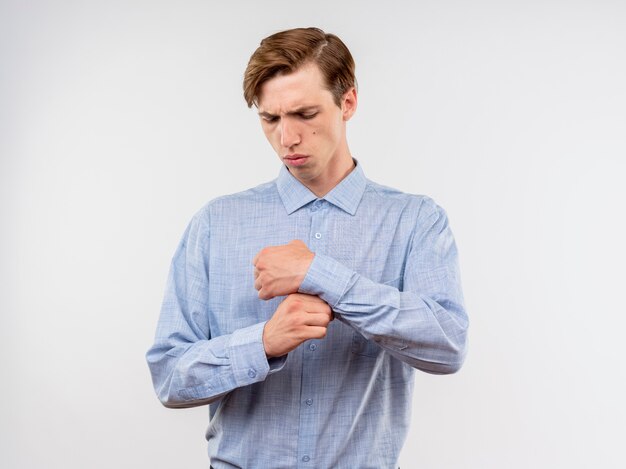 Jeune homme en chemise bleue regardant vers le bas avec un visage sérieux finxing ses boutons de manchette debout sur un mur blanc