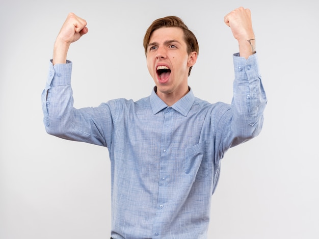 Jeune homme en chemise bleue serrant les poings heureux et excité se réjouissant de son succès debout sur un mur blanc