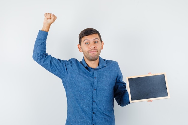 Jeune homme en chemise bleue tenant un tableau noir avec un geste de gagnant et l'air heureux, vue de face.